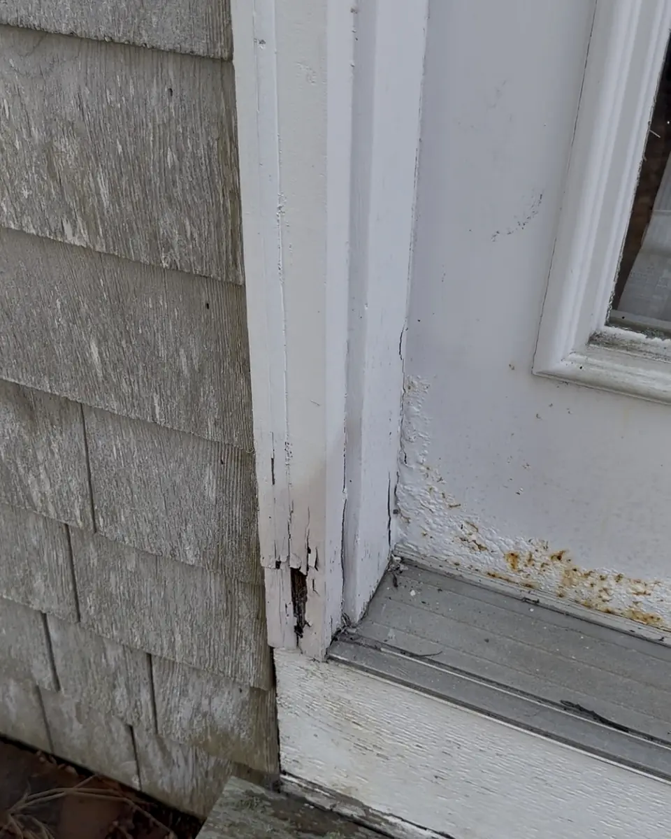 White door trim rotted and split at the bottom, next to weathered cedar shingles and a door sill